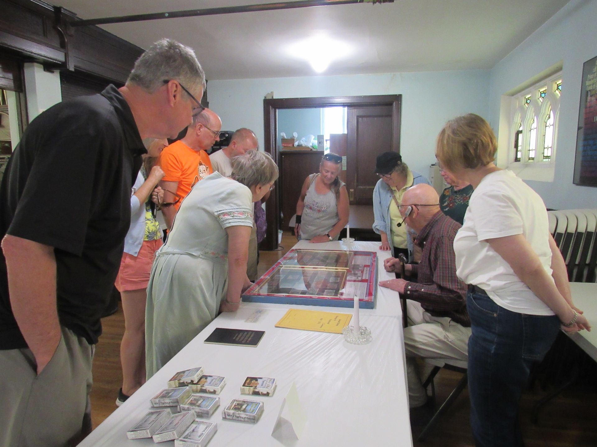 People gather around a table, examining items. Some look at a display case; others watch. Indoors, with a long white table.