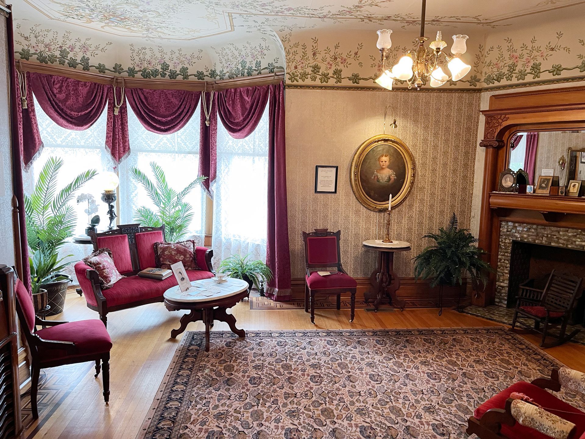 Victorian-era parlor: red velvet sofa and chairs, bay window with curtains, ornate fireplace, patterned rug, wallpaper.