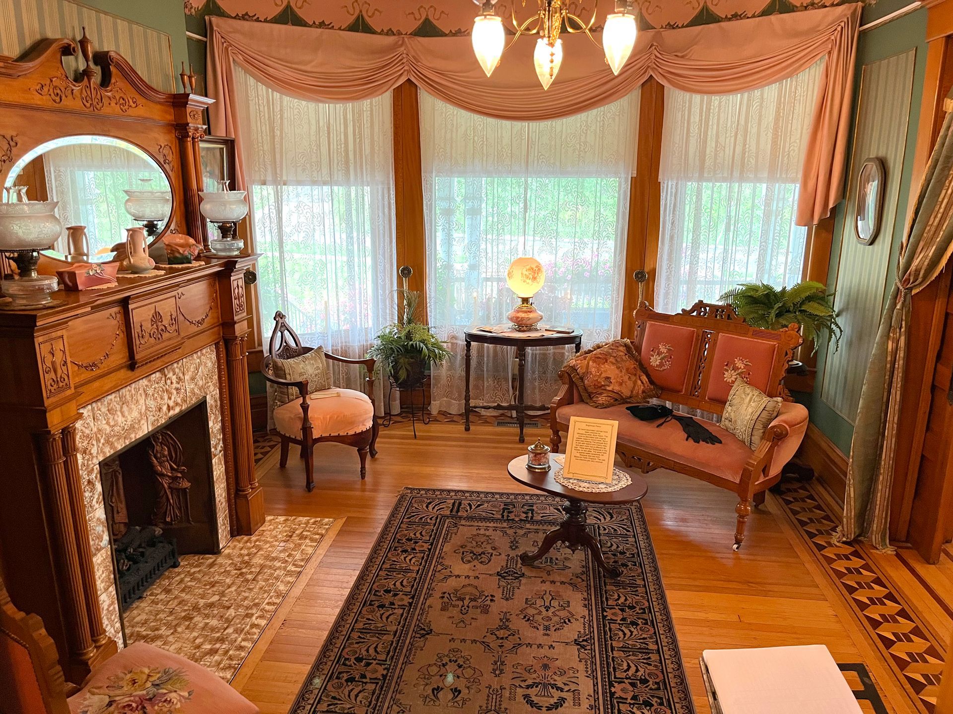 Victorian-era parlor with fireplace, floral accents, pink drapes, and antique furniture.