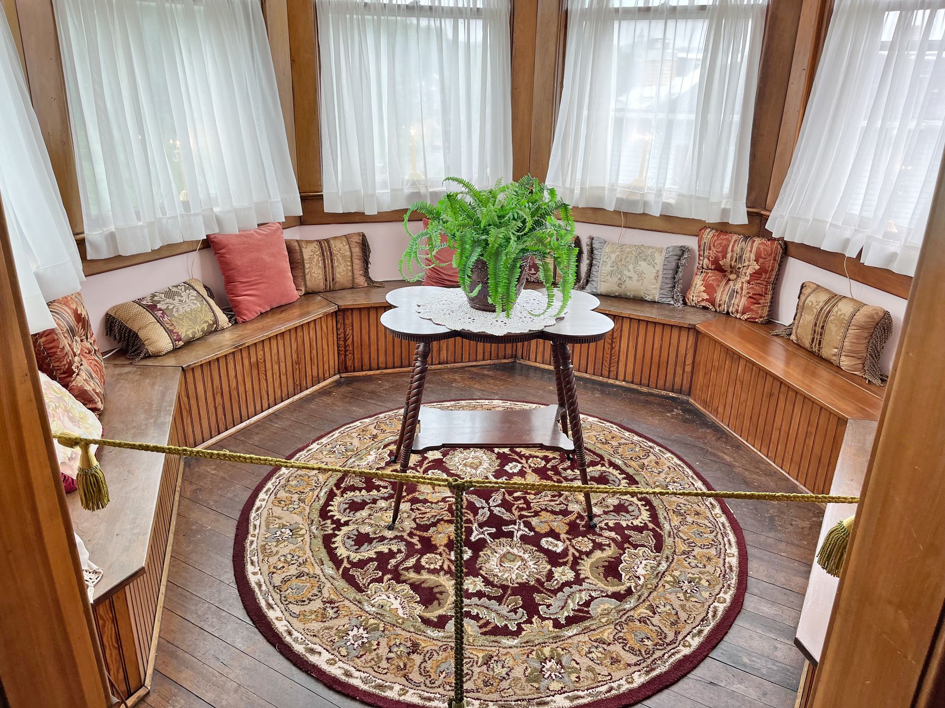 A cozy sunroom with built-in seating, round rug, small table with plant, and sheer curtains.