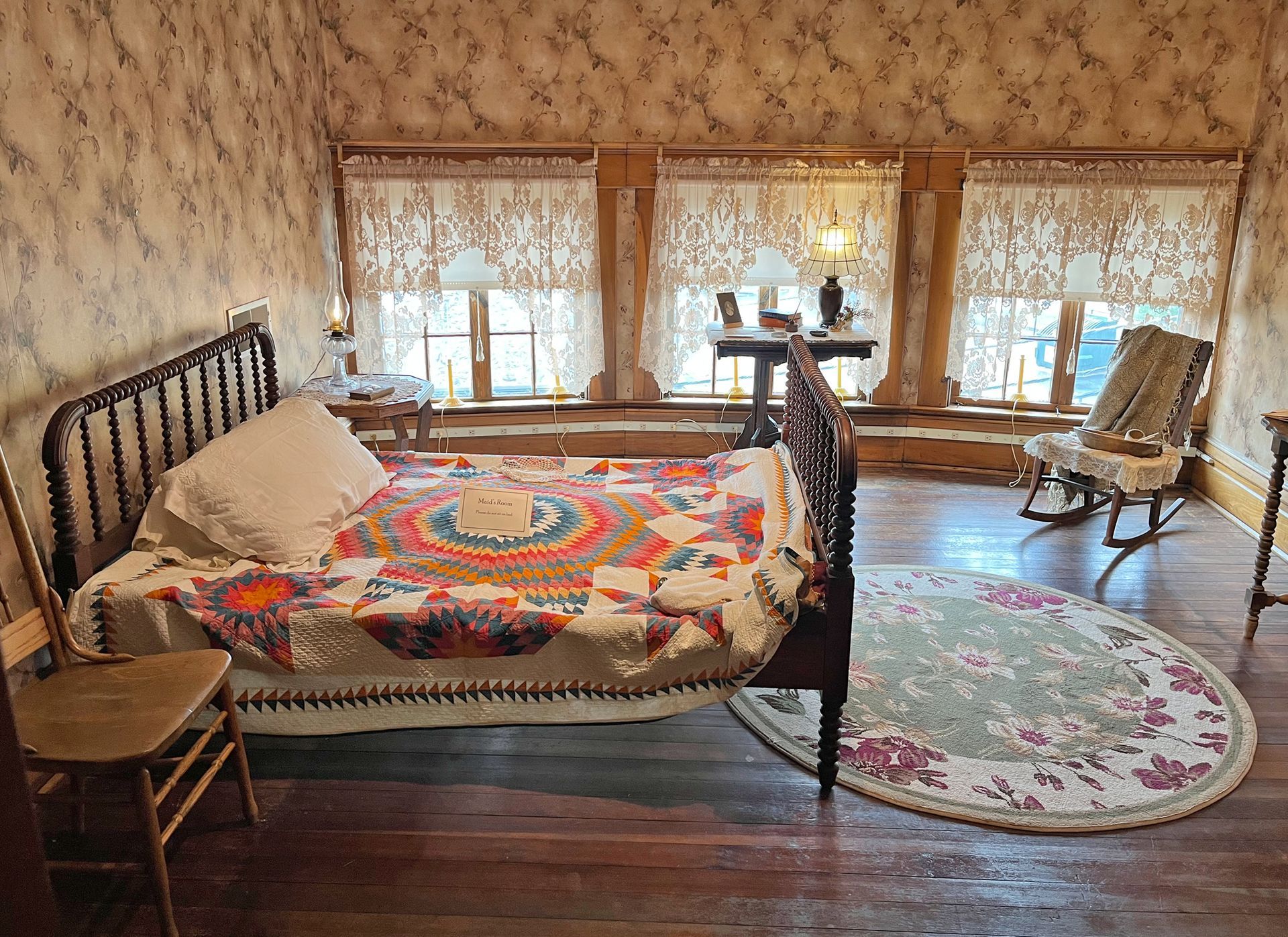 Bedroom with bed, quilt, round rug, and rocking chair. Wallpaper, curtains, and lamps.