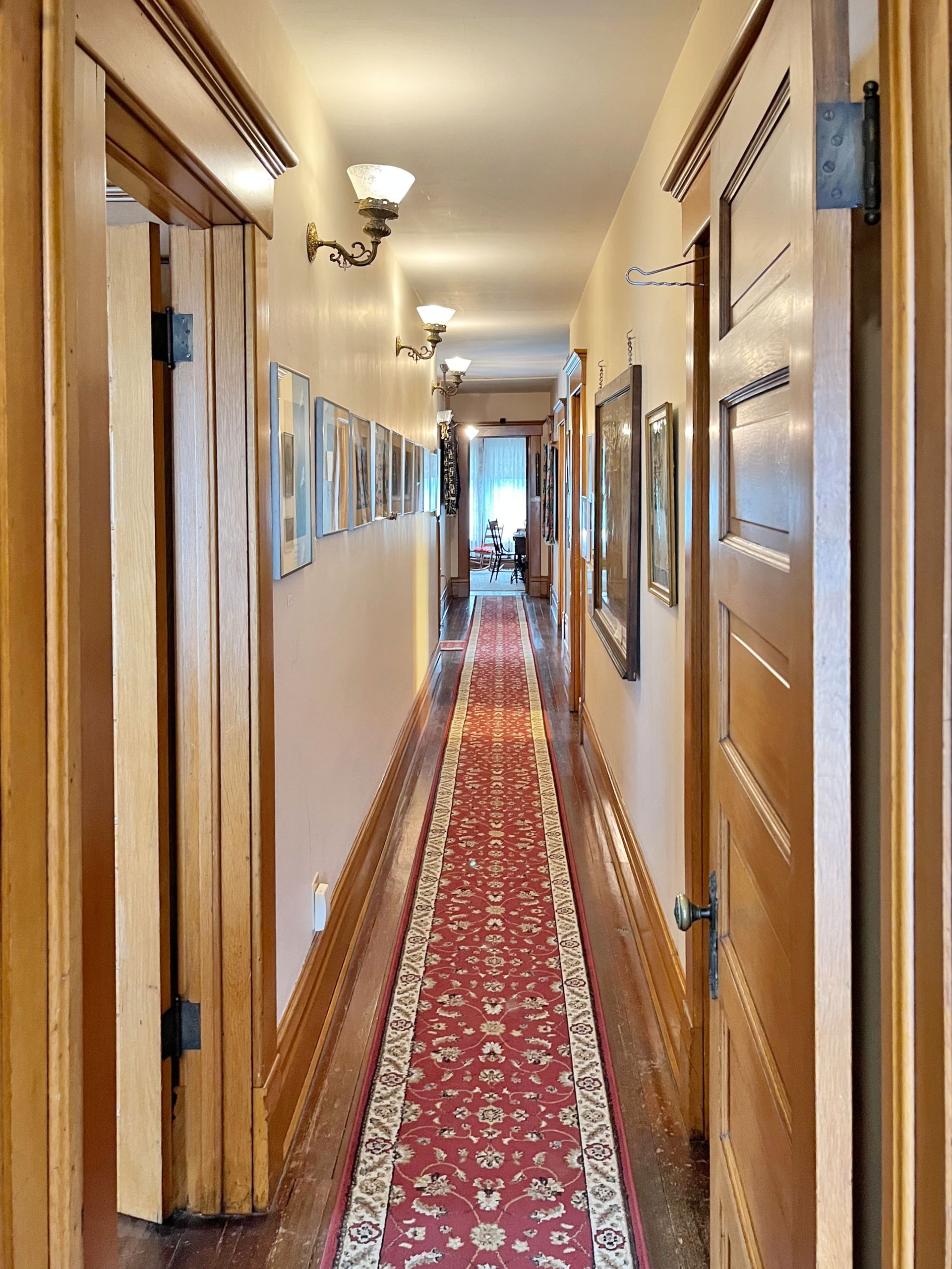 Long, narrow hallway with red runner rug, wooden doors, and framed artwork.