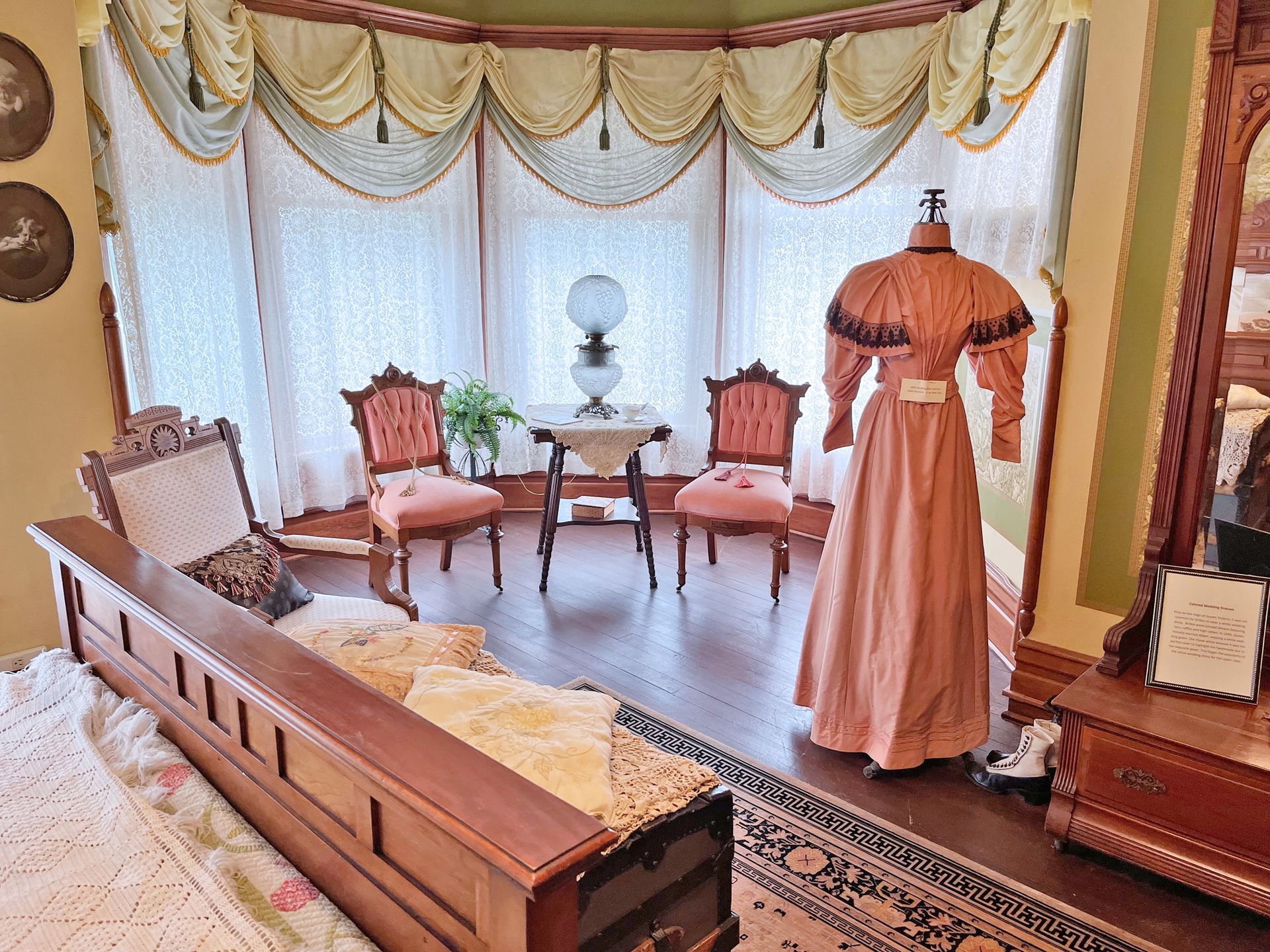 Victorian-era room with a gown on a mannequin, antique furniture, and a bay window.