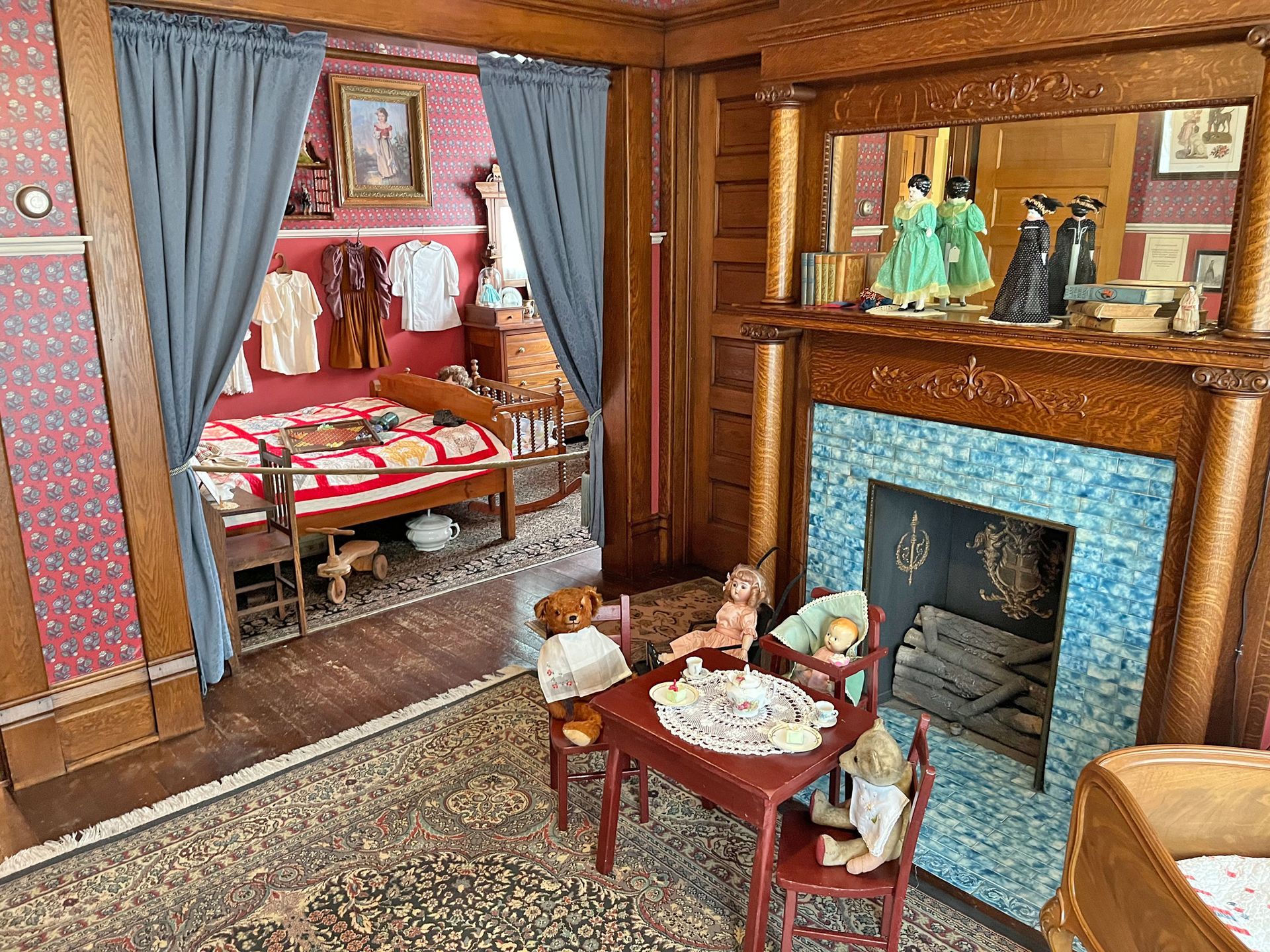 Room with dolls, miniature furniture, fireplace, and red patterned wallpaper.
