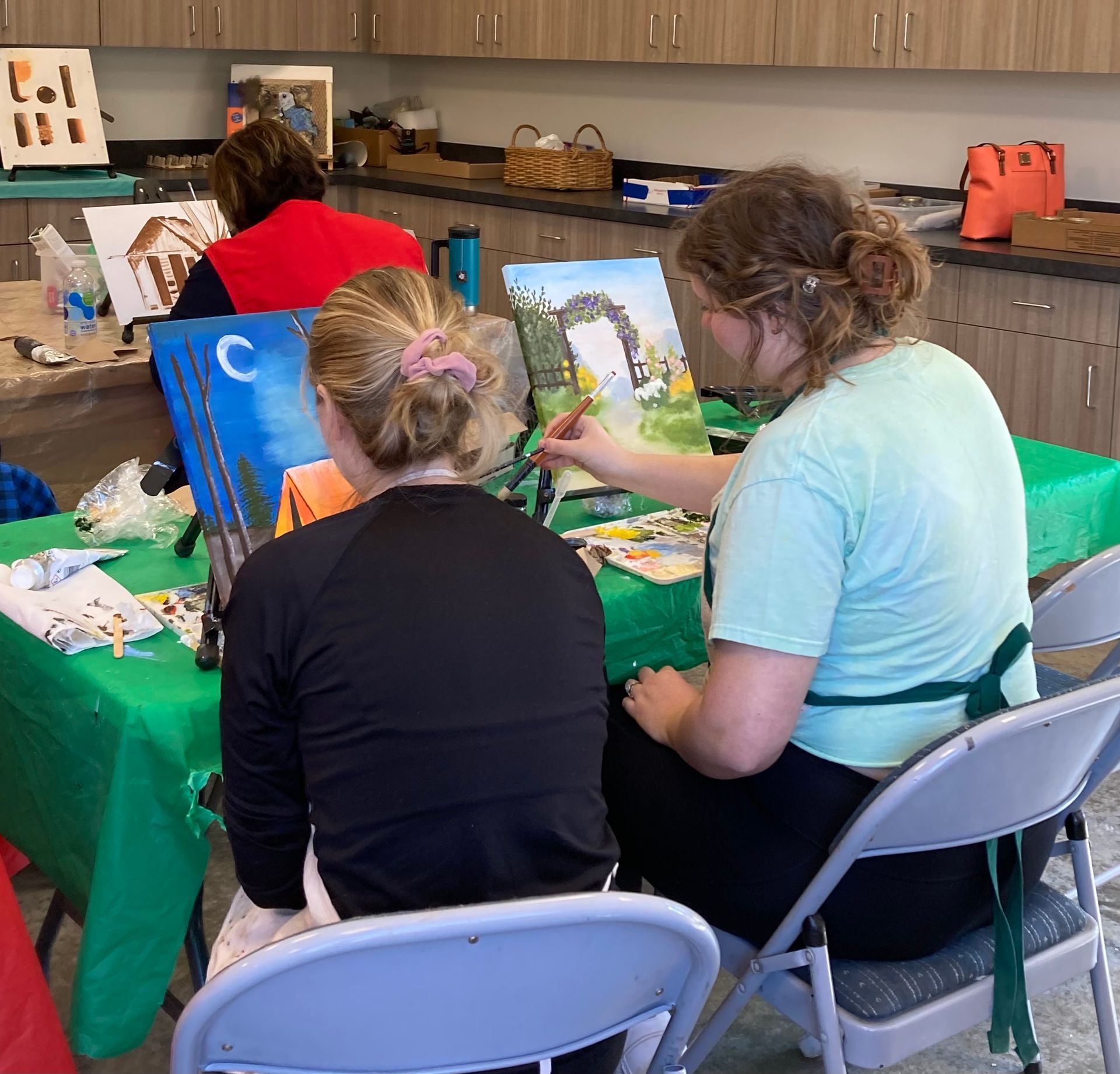 People painting at a table covered in green fabric. One person points to a painting, helping another.
