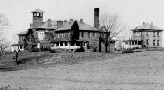 Large brick building with a tower, chimney, and smaller building to the right, on a grassy hill.