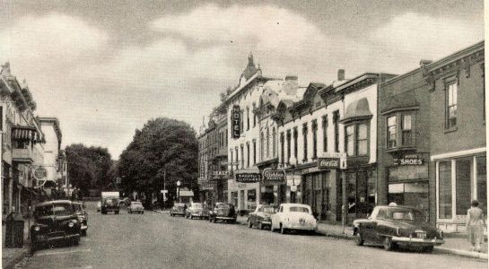 Street scene with cars parked along a row of brick buildings; several storefronts visible, trees along the street.