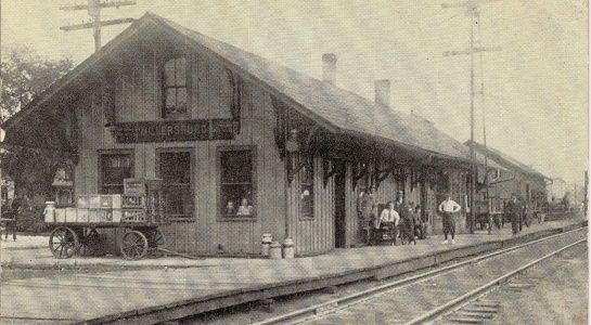 Old wooden train station with people standing on a platform, railroad tracks in foreground.
