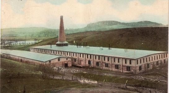 Factory with tall chimney, situated in a rural landscape with rolling hills.