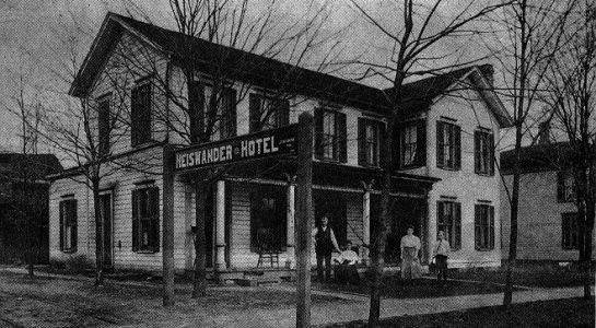 Old black and white photo of the Rosenader Hotel with a sign. Two-story building, porch, and people standing outside.