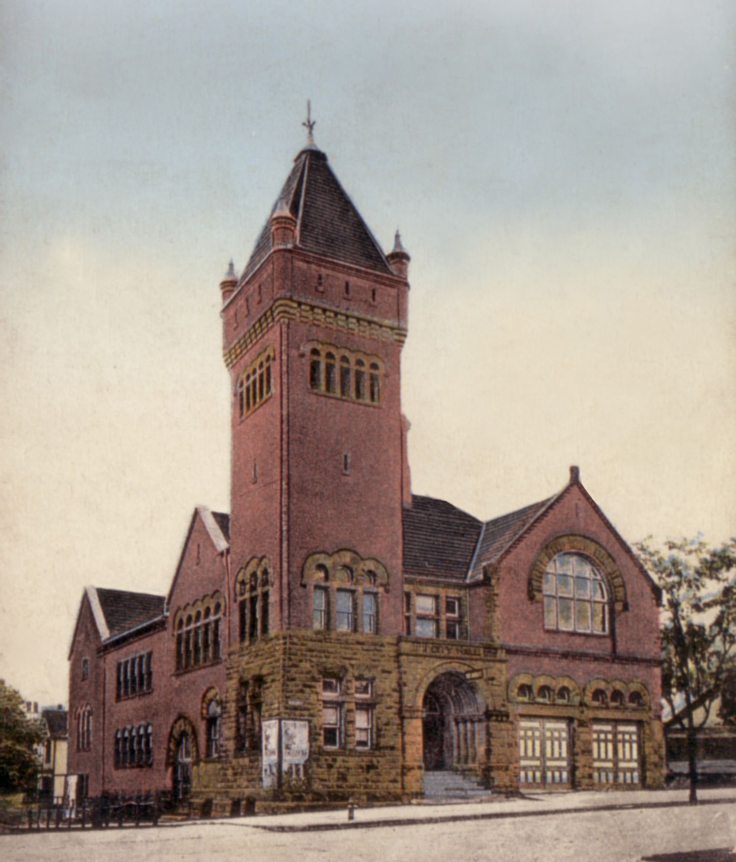 Red brick building with a tall tower, arched entryway, and multiple windows on a city street.