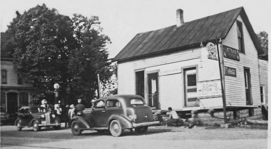 Vintage black and white photo of a small shop with cars parked in front, people gathered near the entrance.