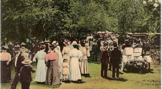 Crowd gathered outdoors, many in early 1900s attire. Automobiles are visible. Green trees and grass form the background.