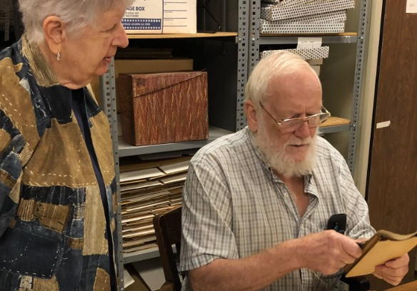 Man with beard examining document, woman watches. Shelves with boxes in background.