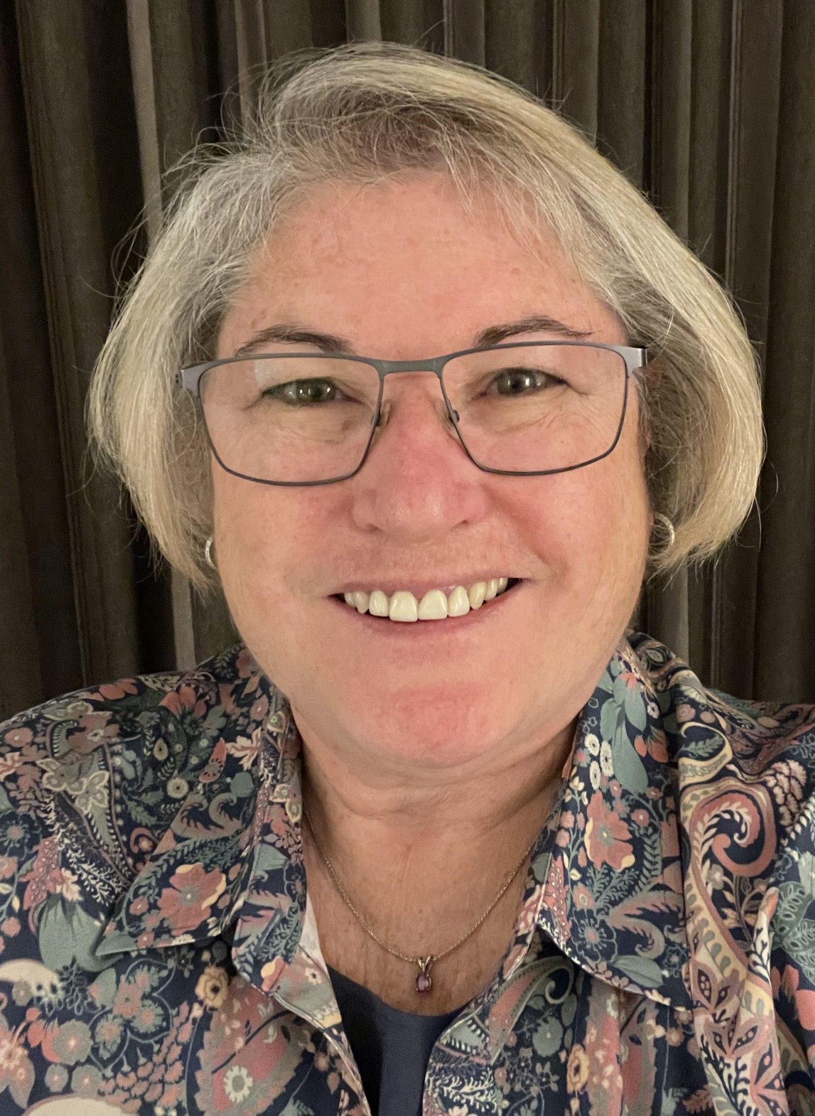 Woman with glasses smiles; wearing patterned shirt, short gray hair.