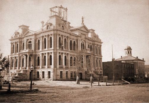 A large, ornate courthouse with a cupola, alongside a smaller domed building. Sepia tone.