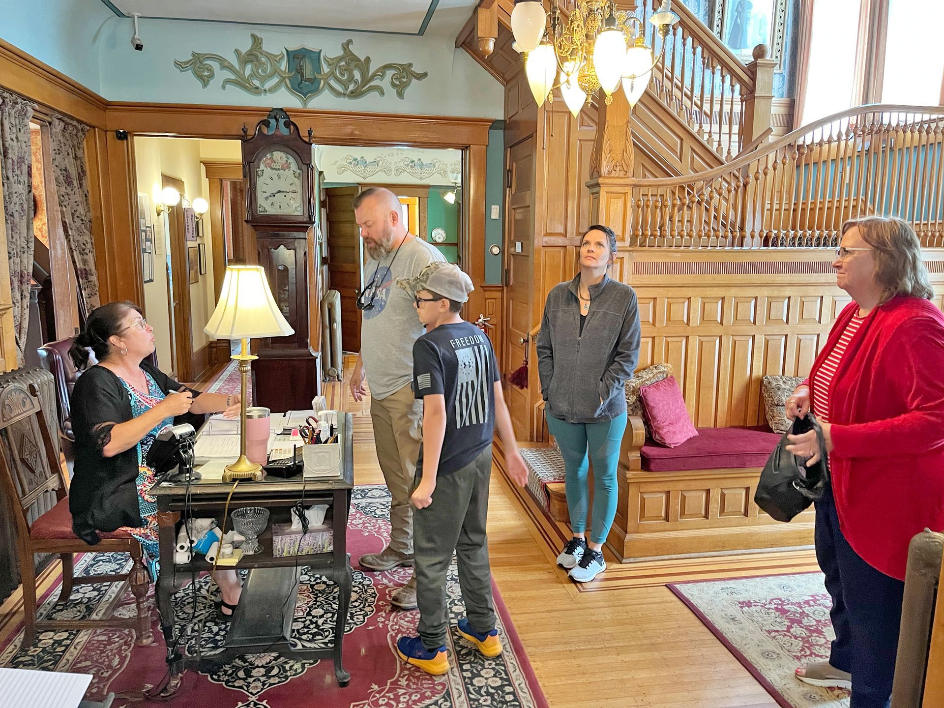 A tour group listens to a woman speaking in a richly decorated room with ornate wood and a grandfather clock.