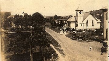Dirt road in a small town. Church with steeple, buildings, and people. Trees line the road.