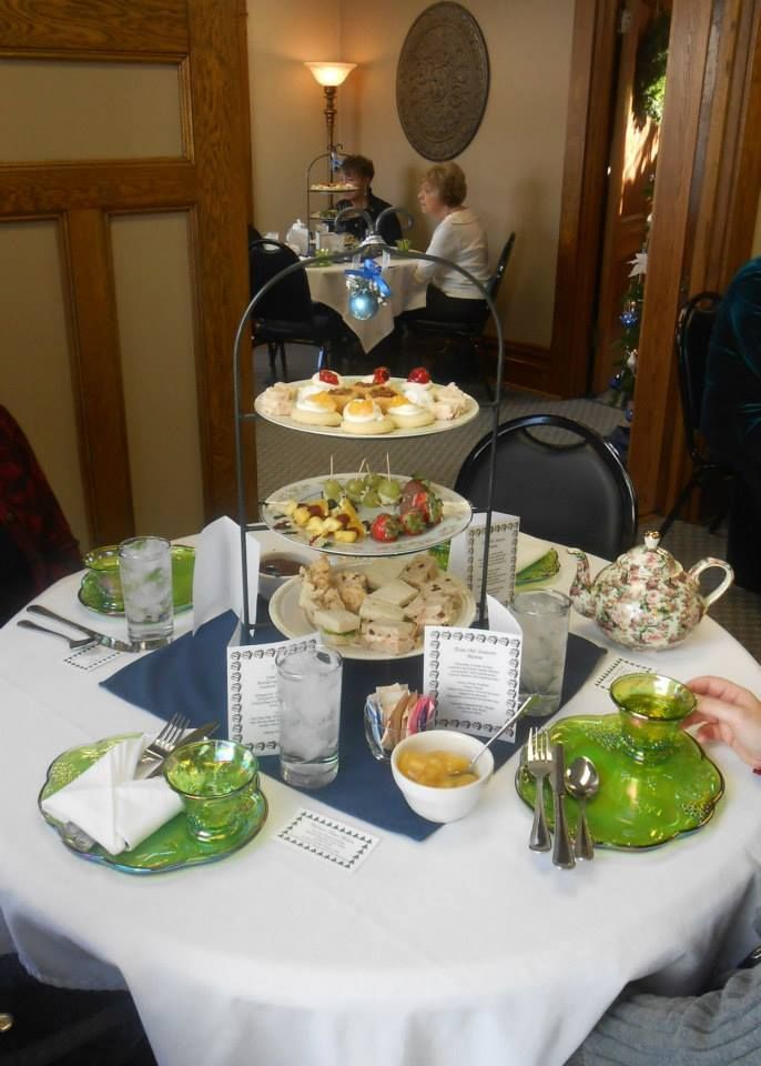 Table set for tea with a tiered tray of food. Two people sit at a table in the background.