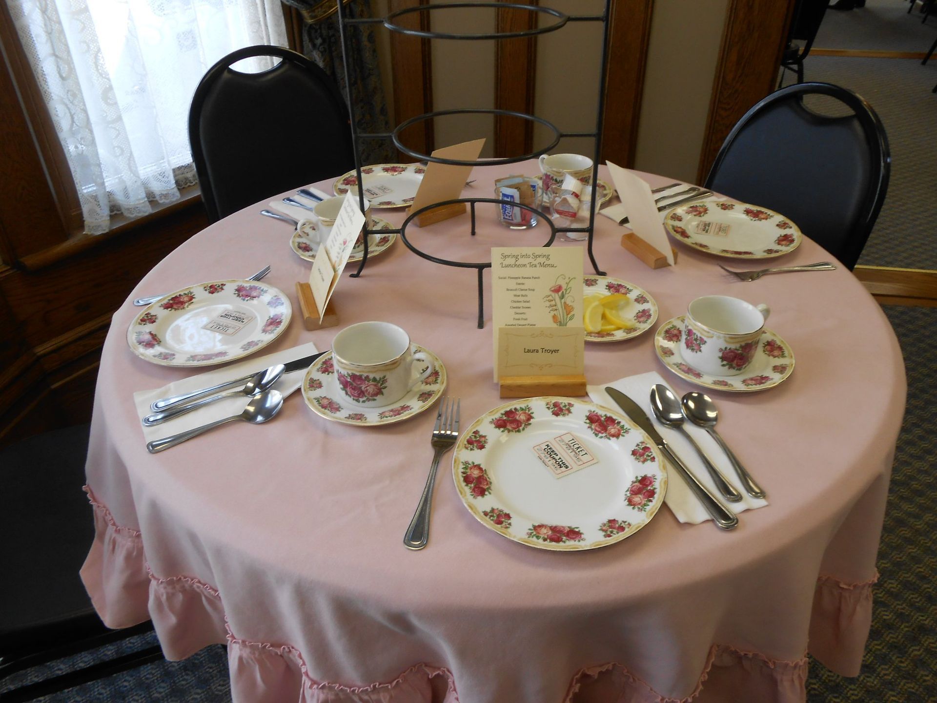 Round table set for tea with pink tablecloth, floral plates, and tiered serving stand.