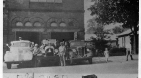 Fire station with three fire trucks, people in front, a child, and a nearby building.