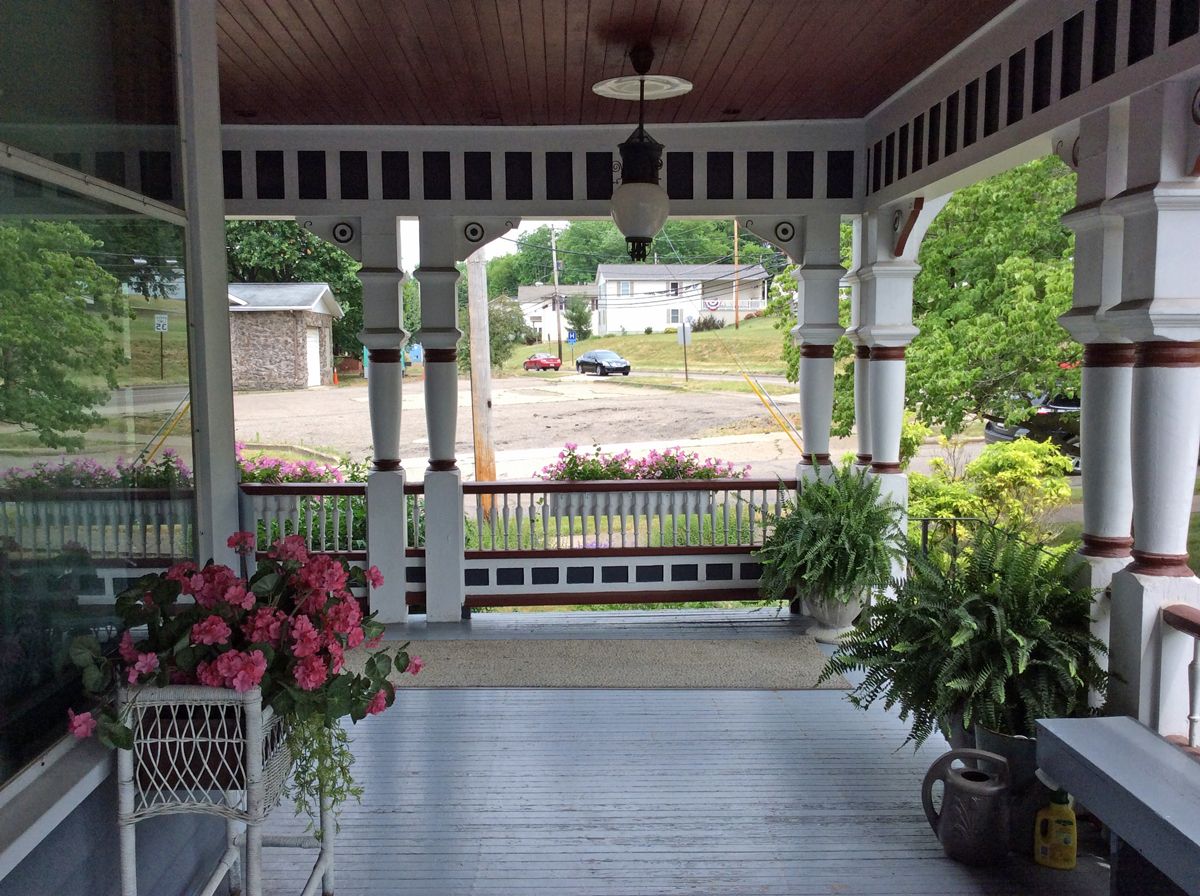 Porch of a white Victorian home with flower pots, overlooking a yard and distant buildings.