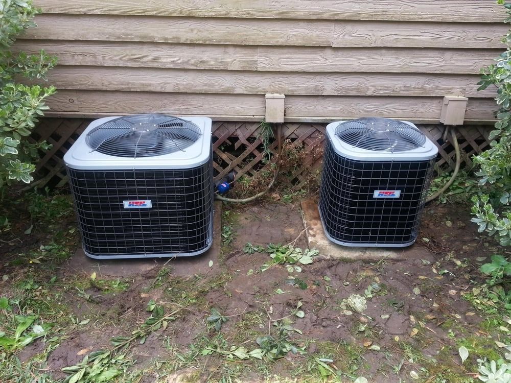 Two outdoor air conditioning units beside a house wall on a patchy dirt yard