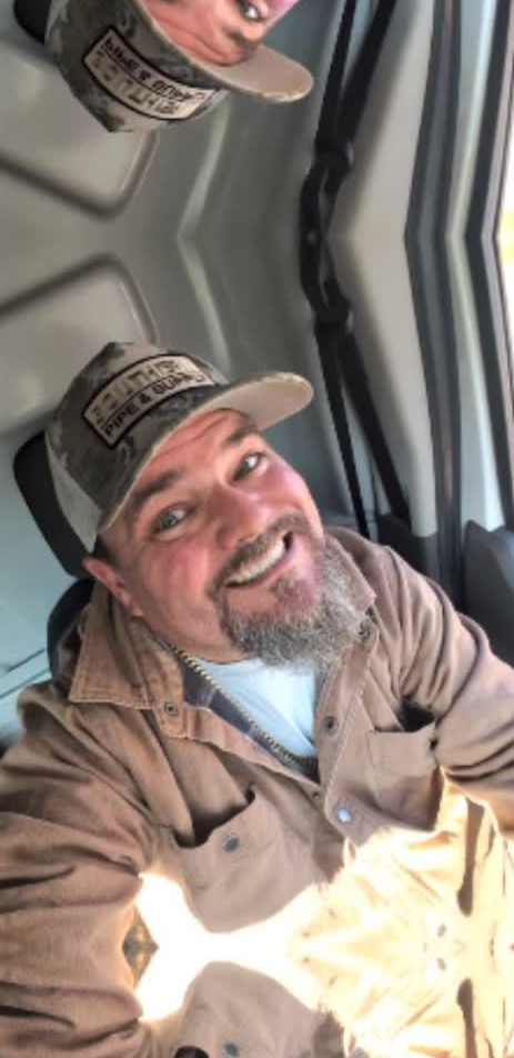 Smiling person in a tan cap and jacket inside a vehicle, with a mirrored reflection above.
