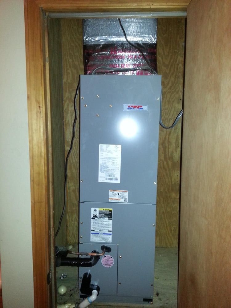 Gray HVAC furnace in a narrow closet with exposed ductwork and pipes.