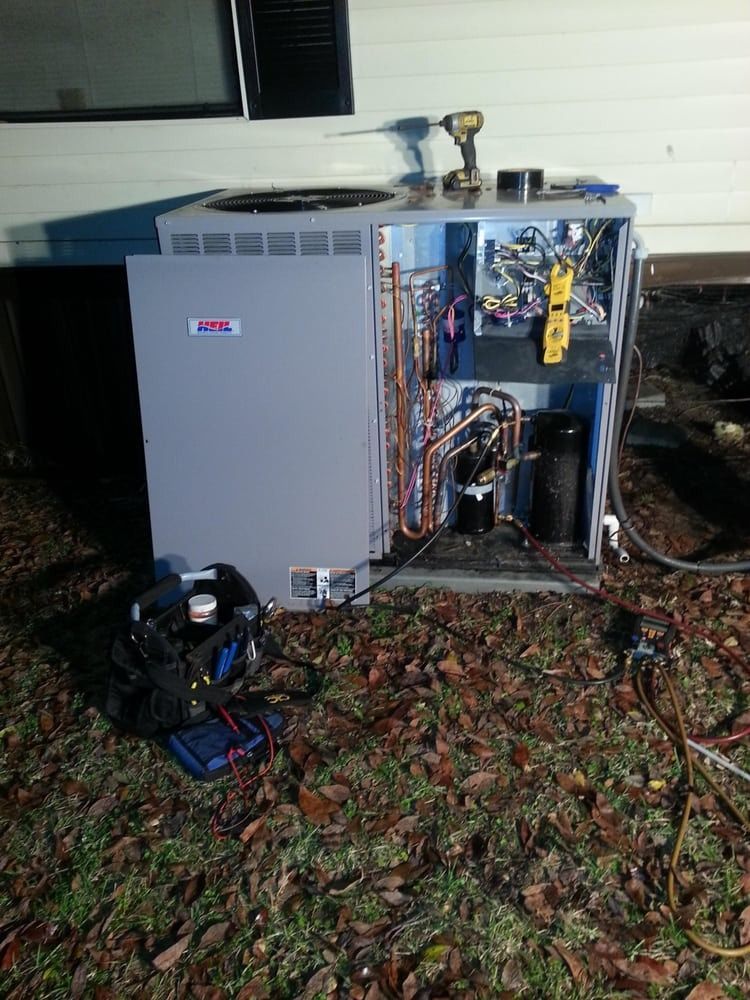 Outdoor HVAC unit with front panel open, exposing wiring and components beside a house at night.