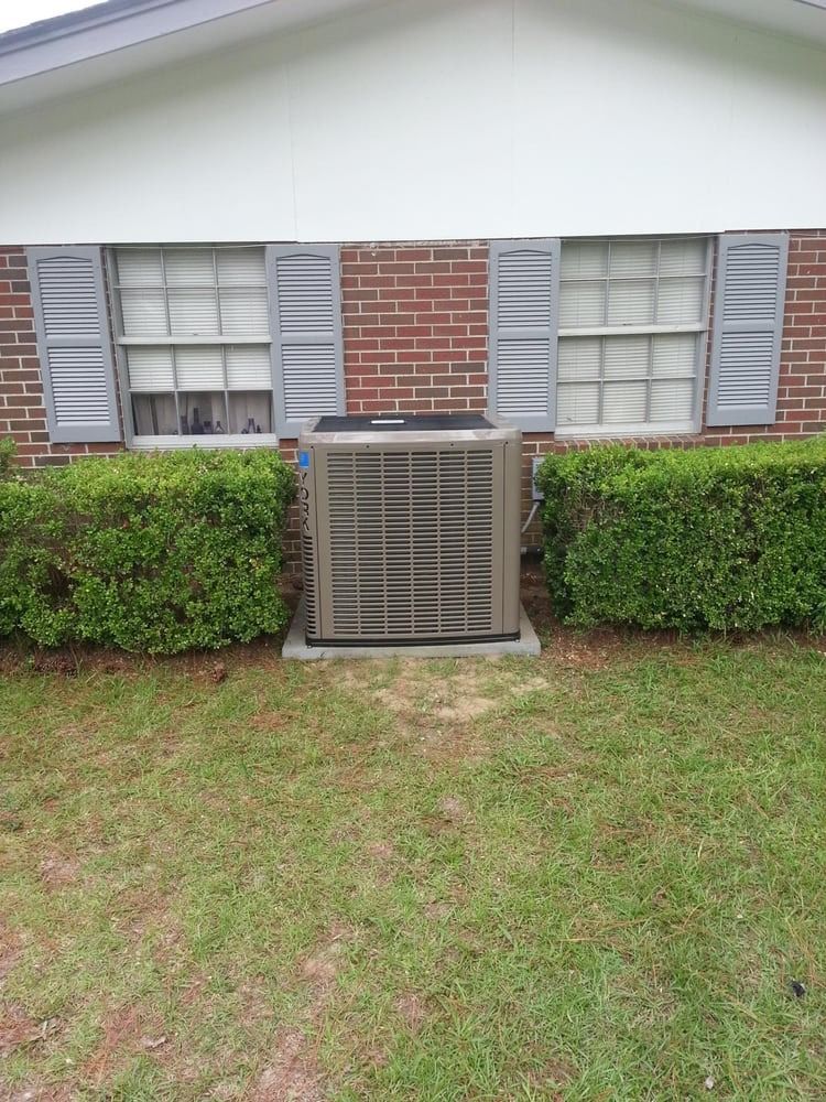 Residential building exterior with a central HVAC unit, brick wall, windows, shutters, hedges, and grass lawn