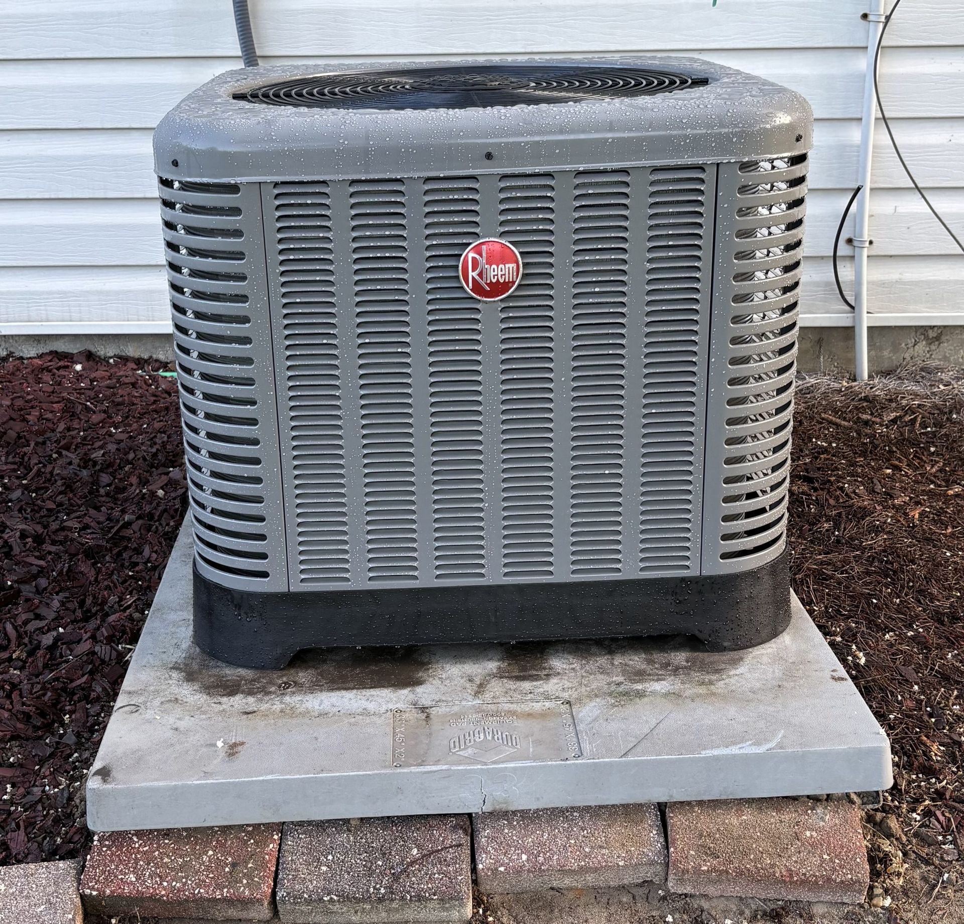 Outdoor gray HVAC unit on a concrete pad beside a house, with a red logo on the front
