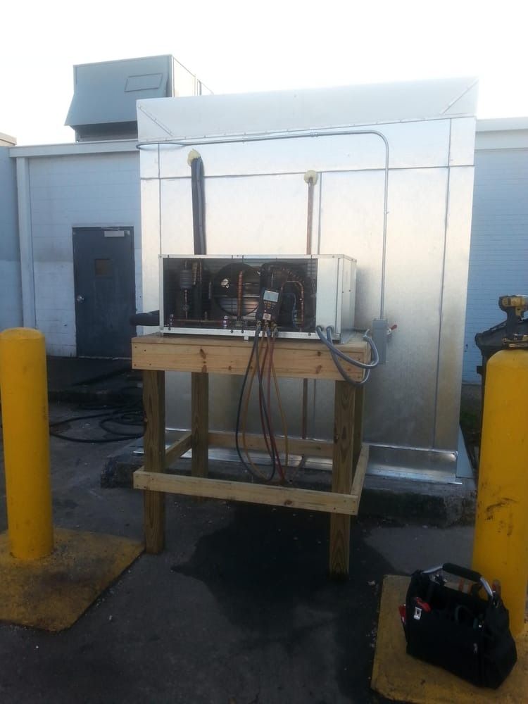 Industrial machine on a wooden stand in a parking lot beside a white building, with yellow bollards in front.