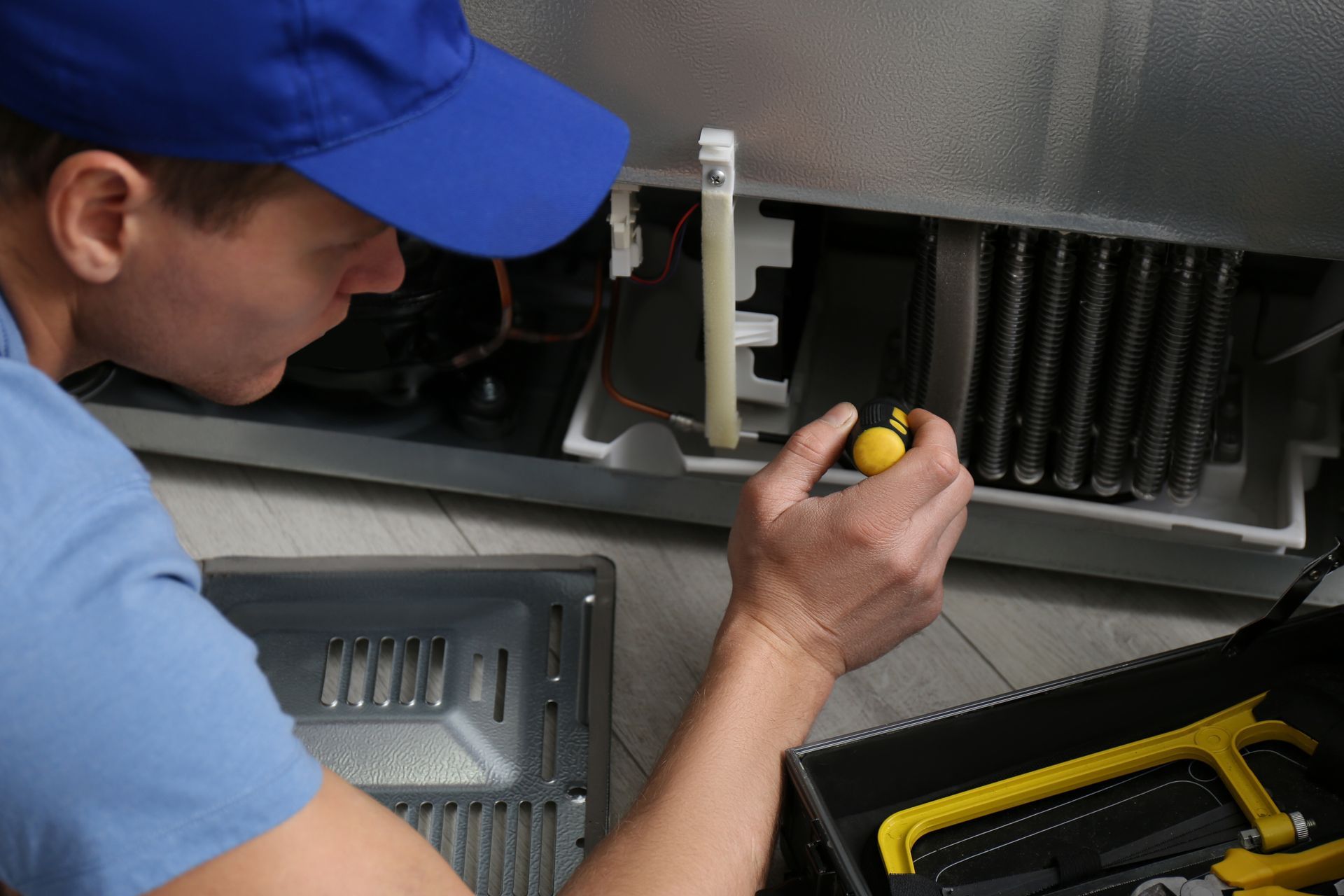 Technician in blue cap uses a screwdriver inside an open appliance with tools nearby