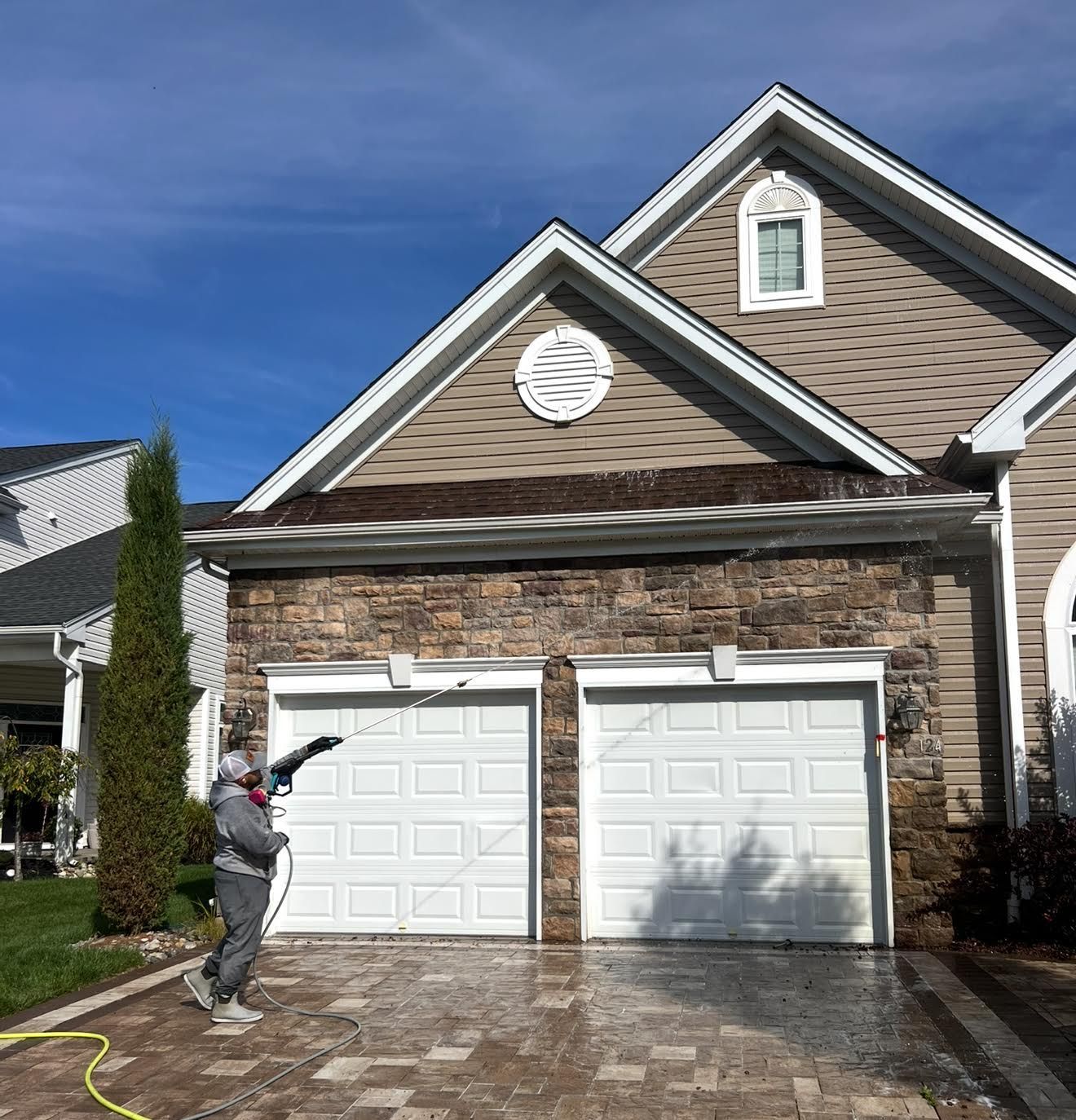 Person spraying a two-car garage on a brick suburban house under a blue sky
