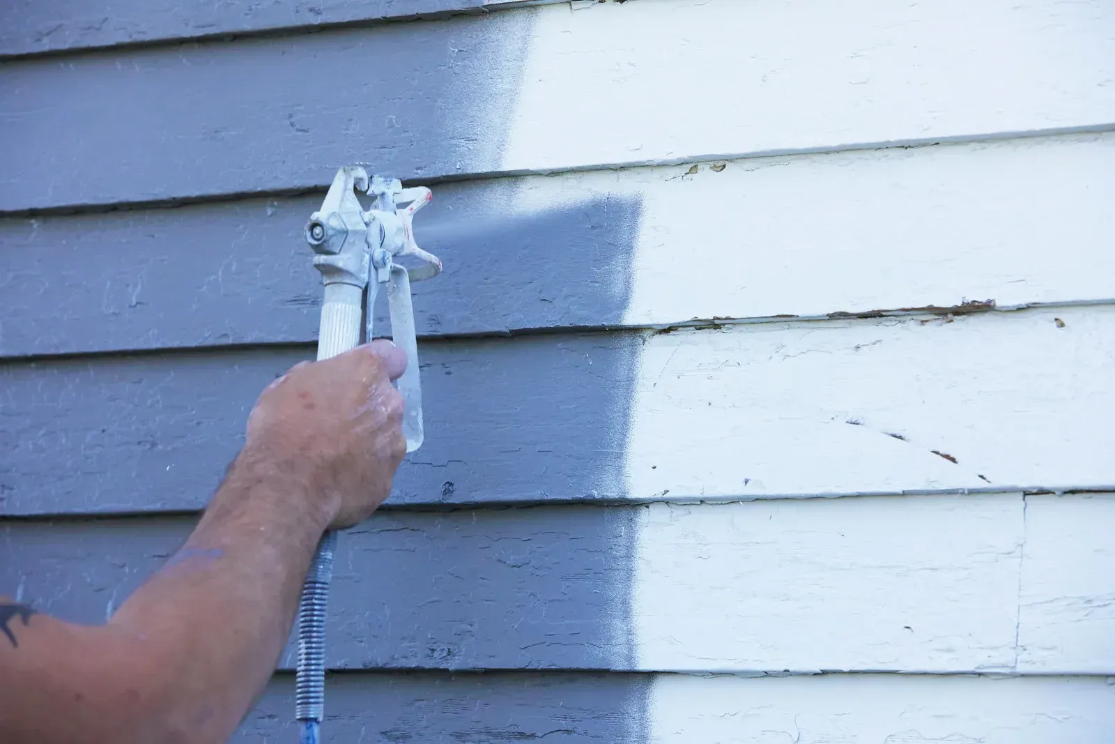 Hand spraying gray paint onto white house siding during exterior painting