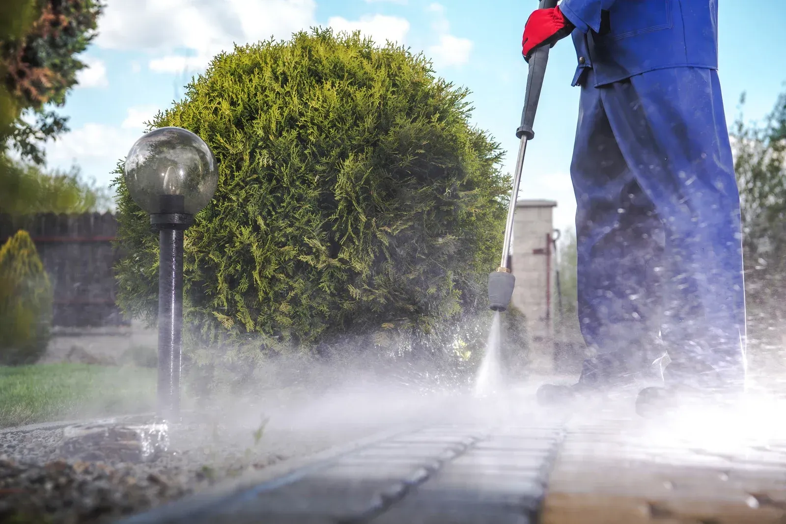 Person pressure washing a stone walkway beside a trimmed hedge and garden light.