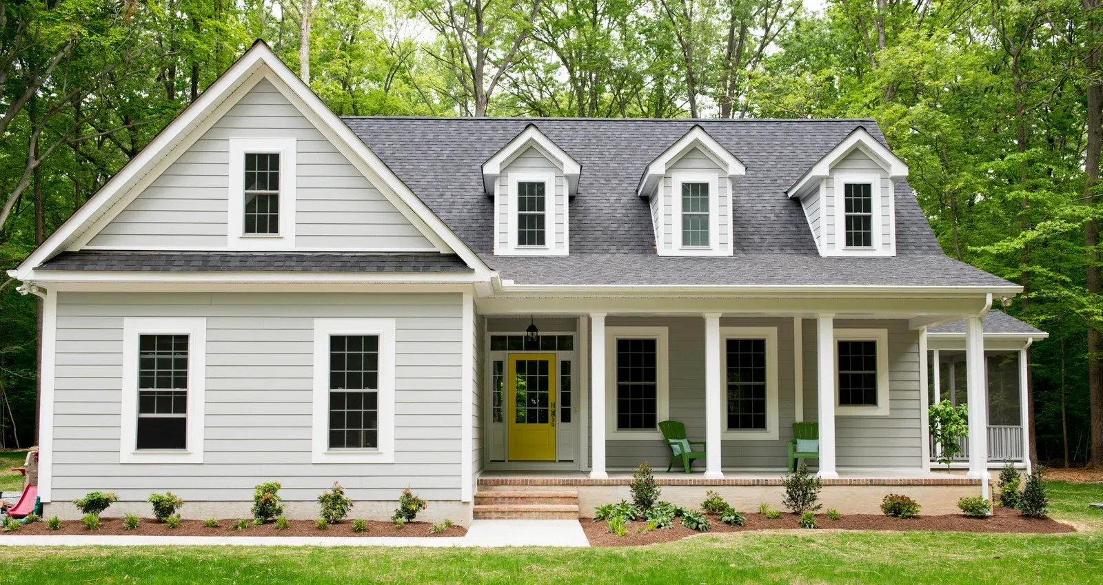 White suburban house with a gray roof, yellow front door, and porch surrounded by green trees