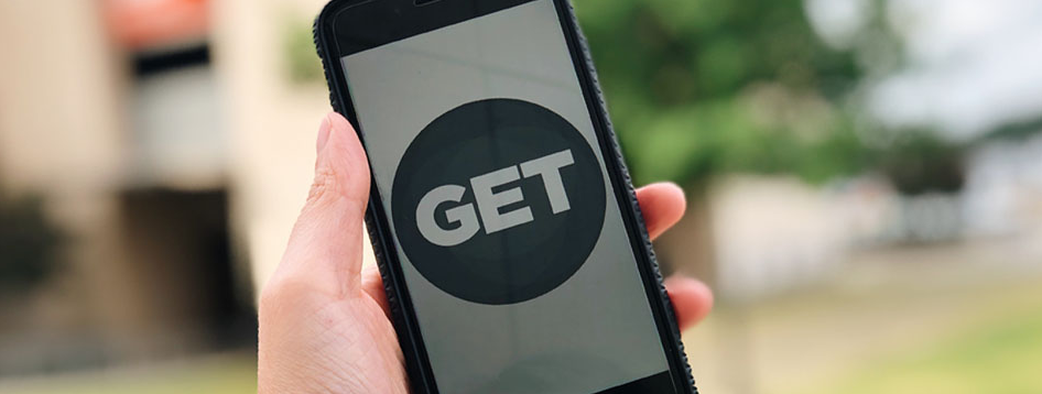 Hand holding a phone with a Transact logo on screen, set against green foliage. The phone is in a black case.