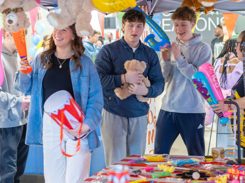 Three people holding prizes like stuffed bears and a drum at an outdoor event.