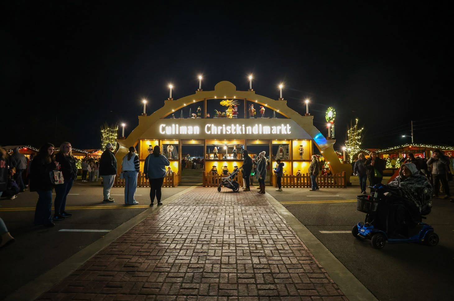 Nighttime entrance under the largest walk through Schwibbogen at the entrance of the Cullman Christkindlmarkt, people walking, and cobblestone path