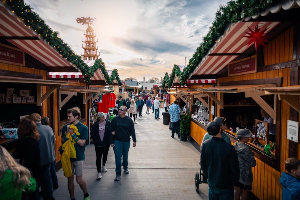 Open-air market aisle with people walking between wooden stalls under striped awnings, at sunset