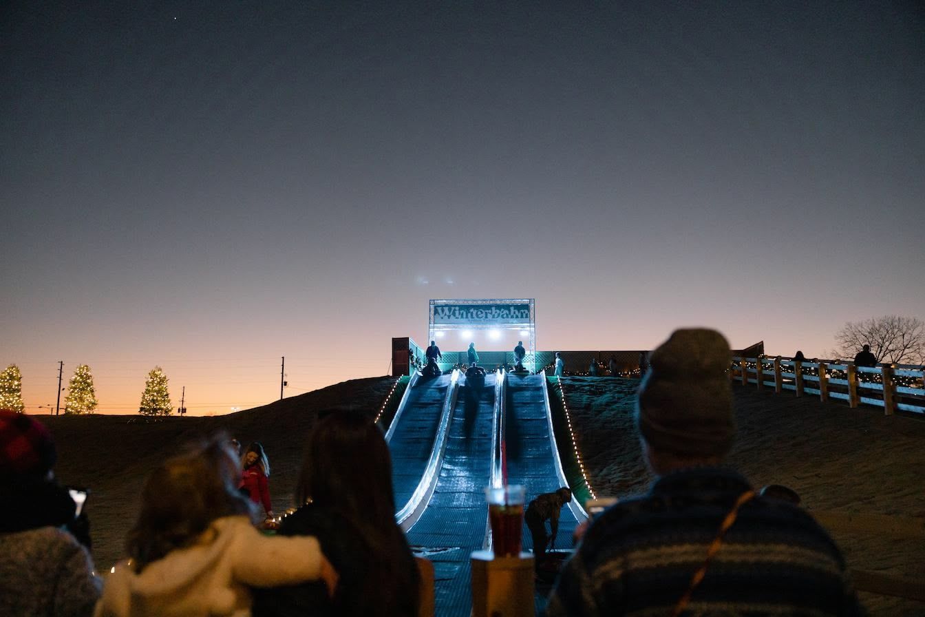 People watch a long, illuminated tubing slide at night. Blue lights line the path, leading to a bright structure.