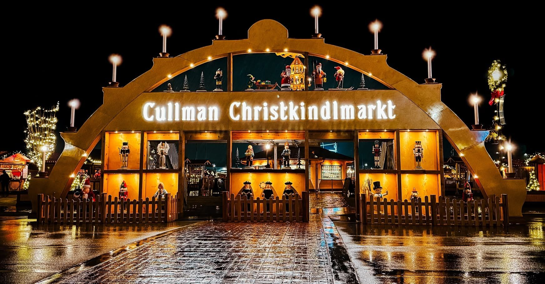 Cullman Christkindlmarkt lit up at night, with glowing orange arches and reflections on wet pavement