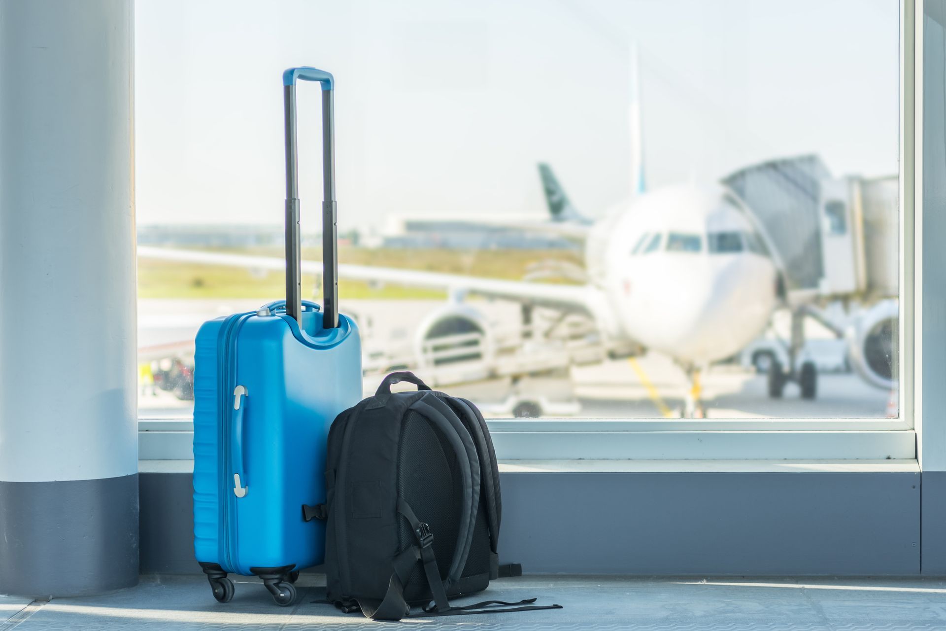 Una maleta azul y una mochila negra están frente a una ventana en un aeropuerto.