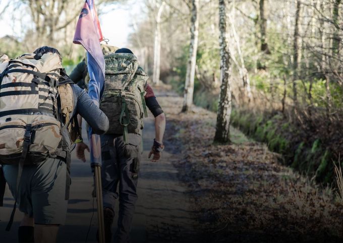 Twee wandelaars met rugzakken lopen over een onverhard pad in het bos, waarvan er één een vlag draagt.