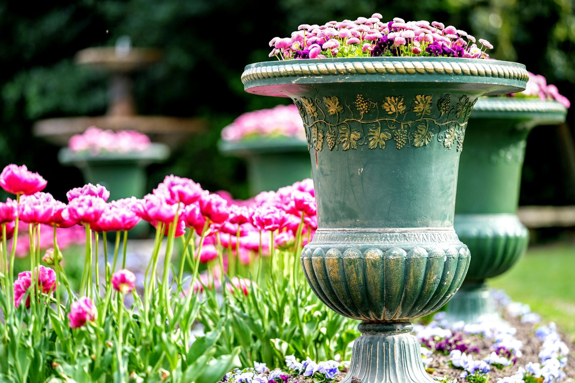 Pink flowers in a garden beside ornate green urn planters with blooming flowers on top