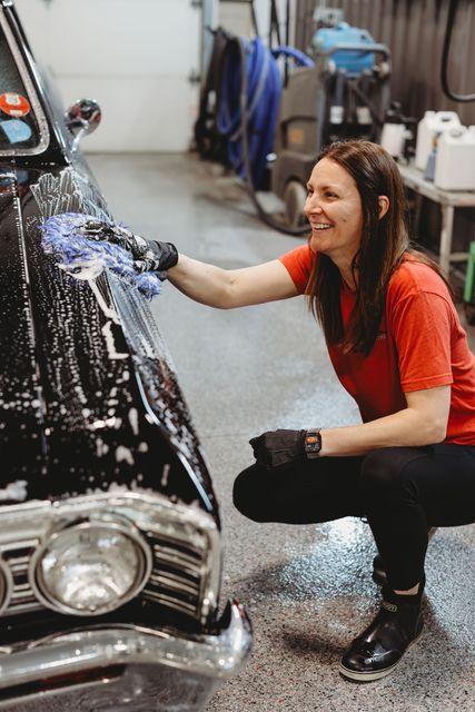 Person washing a black car in a garage, smiling and crouching beside the hood.