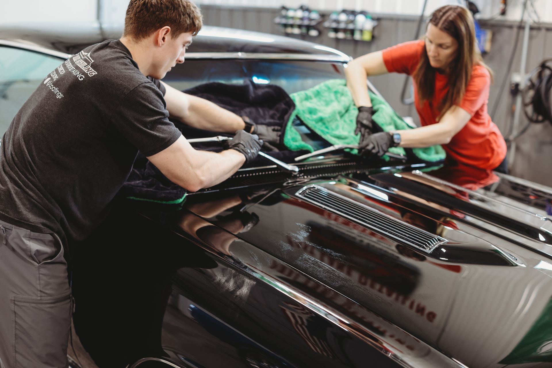 Two people cleaning a black car with cloths and spray in an auto detailing shop