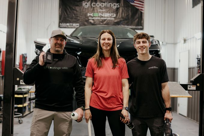 Three people standing in a car detailing shop, smiling in front of a black vehicle and garage equipment